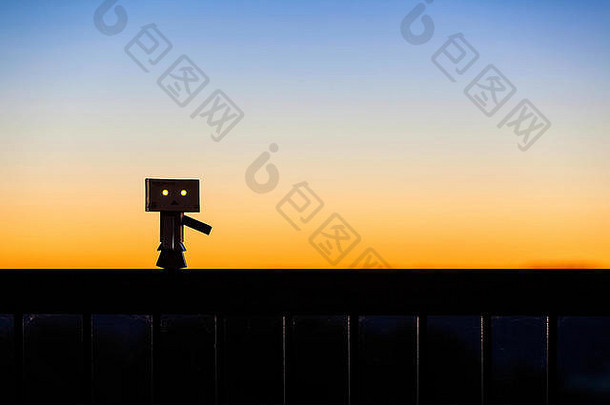 Danbo Danboard虚构的机器人角色在五颜六色的黎明天空衬托下沿着栏杆行走