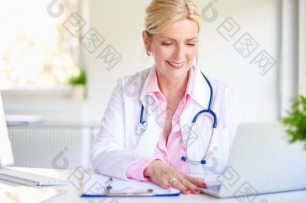 这张特写照片是一位上了年纪的女医生坐在医生办公室的办公桌旁，微笑着用笔记本电脑写东西。