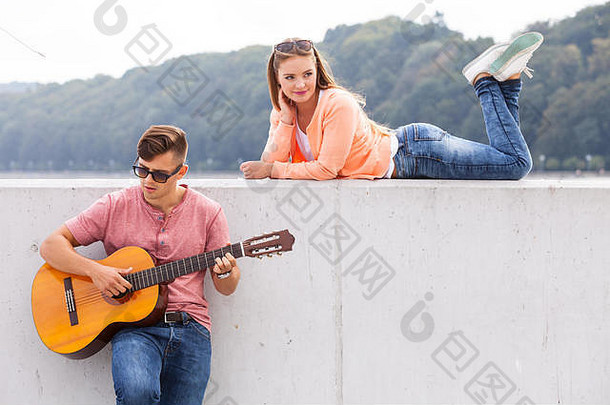 爱情<strong>浪漫音乐</strong>天才激情约会概念。吉他手和他的缪斯。年轻人弹吉他，女孩躺在有风景背景的墙上。