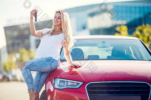 夏日，一位穿着牛仔裤、手里拿着钥匙的快乐金发女郎站在红色汽车旁的照片