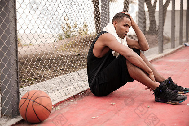 疲惫的非洲<strong>篮球</strong>运动员拿着毛巾坐在<strong>篮球</strong>场附近的公园里的照片。往旁边看。用手握住他的头。