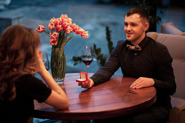 年轻男子向女子求婚，并为她提供带订婚戒指的首饰盒。他们坐在桌子旁，旁边是几杯酒和花瓶