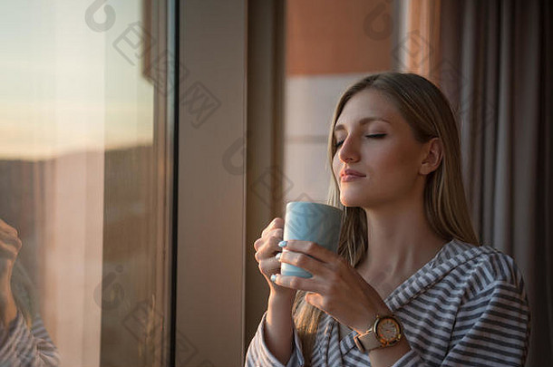 身穿浴袍的年轻女子站在窗前，享受着傍晚的咖啡和城市美丽的日落景色