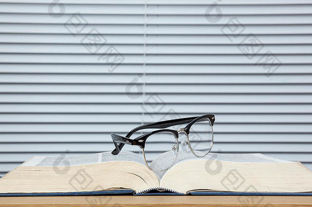 一副黑框眼镜放在一本破旧的打开的教科书上，背景是百叶窗