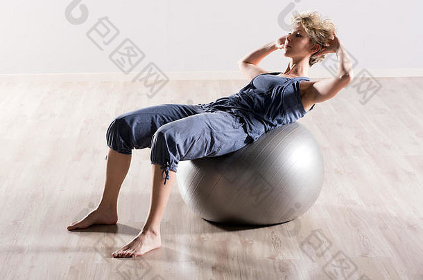 在硬木地板上，一位年轻的赤脚女士双手放在灰色的大稳定球上做仰卧起坐