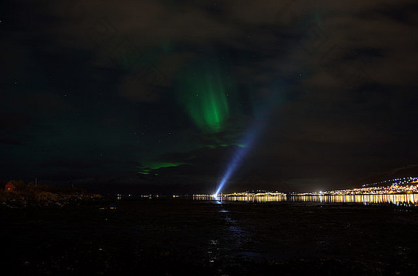 峡湾上的船只探照灯上方的北极光反射着地面，背景中有照明的居民点