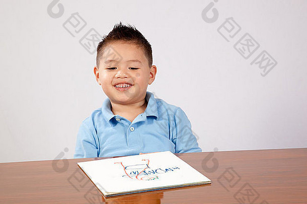 可爱的小男孩坐在木桌旁，手里拿着纸和铅笔，在纸上写字和画画