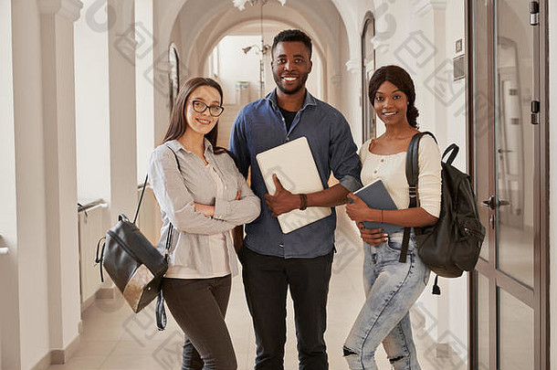 三位非洲和高<strong>加</strong>索人<strong>好友</strong>站在大学走廊的正视图，手里拿着背包和便条。学生们在大学里一起上课。