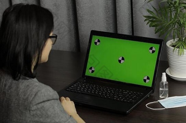 女士在笔记本电脑键盘上有一个视频电话会议，键盘上有绿色屏幕和色度键。远程在线工作。医用口罩，笔记本附近的消毒剂。C2019冠状病毒疾病检疫后视图