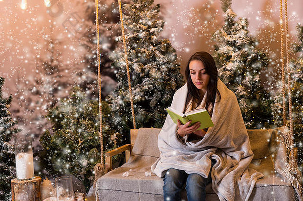 年轻的女人阅读书坐着摇摆不定的包装温暖的毯子白雪覆盖的公园云杉树下雪