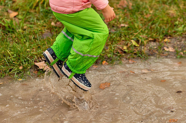 德国十月一个阴雨的秋日，一个穿着绿色防水裤和橡胶靴的小女孩跳进了一个巨大的雨坑