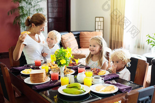 在家吃健康的家庭早餐。母亲和孩子们吃热带水果、烤面包、奶酪和香肠。孩子们在阳光充足的天气喝新鲜的榨果汁
