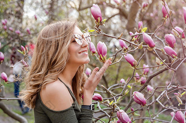 阳光明媚的日子里，春天公园里，一位金发碧眼的年轻女子在盛开的木兰花树旁。木兰树。美丽快乐的女孩在一个房间里享受着香味