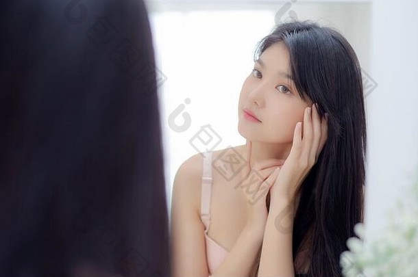 美丽的亚洲年轻女子微笑着看着镜子，用护肤品和化妆品检查面部，以恢复年轻和卫生，美丽的女孩快乐干净