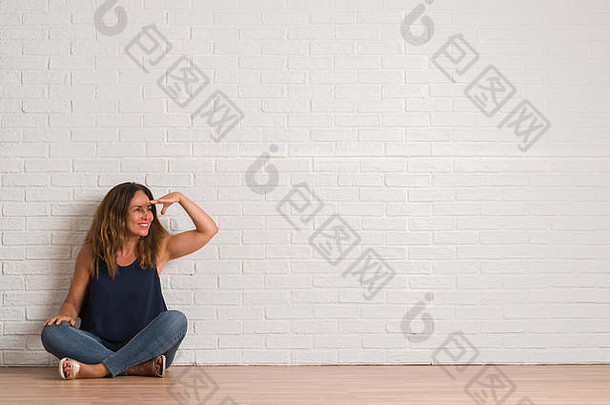 中年西班牙裔妇女坐在白砖墙上方的地板上，非常开心，微笑着用手捂着头远远望去。<strong>搜索</strong>概念。