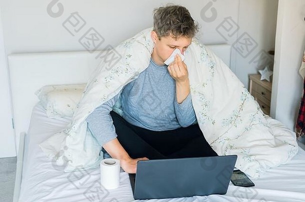 年轻的高加索人生病的男人。吹鼻子坐着床上温暖的毯子工作移动PC工作首页疾病隔离