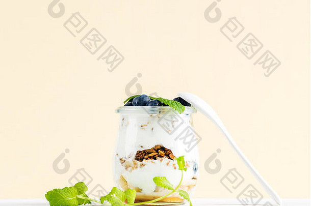 酸奶燕麦格兰诺拉麦片，配果酱、蓝莓和绿叶，置于淡黄色背景下的玻璃罐中。