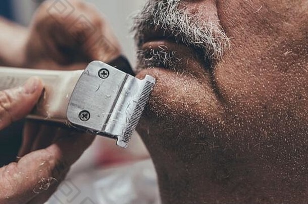 电动剃须刀握在理发师手中的特写镜头。理发师在沙龙里认真地修剪<strong>老客户</strong>的胡须。