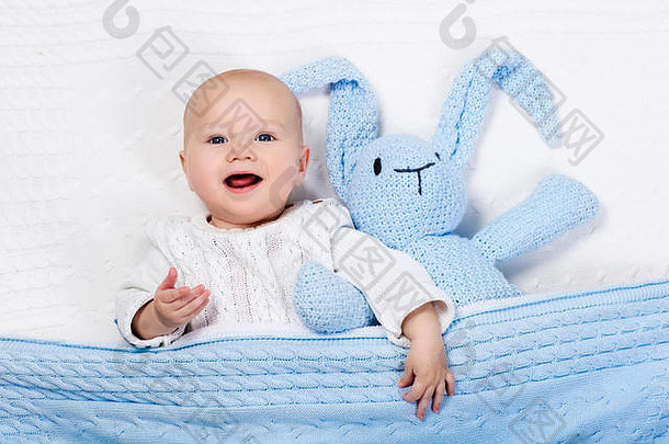在阳光明媚的托儿所里，一个穿着暖和的针织夹克的有趣的小宝宝和玩具兔子在白色电缆编织毯上玩耍