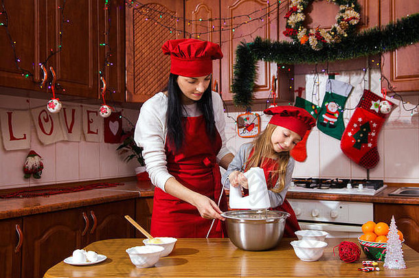 笑容满面的快乐女孩和穿着节日红围裙的母亲在圣诞装饰品前做饭。这家人正在为新年准备甜点。
