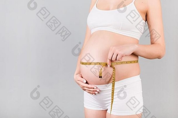 穿着白色内衣的孕妇在灰色背景下测量她正在生长的腹部。英寸卷尺。空间。