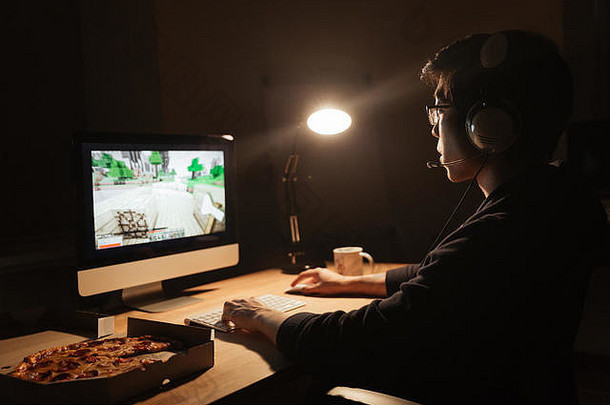 戴着耳机的游戏玩家在黑暗的房间里玩电脑游戏和吃比萨饼