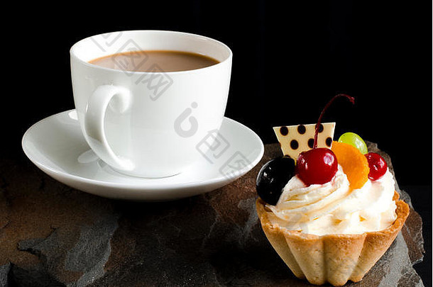 樱桃、葡萄和李子的水果蛋糕站在一块野石头上，旁边是一杯有泡沫的白色咖啡