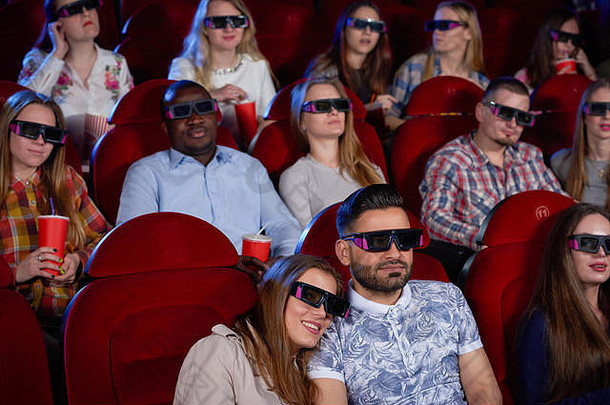 一对阿拉伯男人和一对黑发女人坐在电影院里，拥抱并观看喜剧。帅哥抱着坐在旁边的漂亮女友看电影。现代电影院。