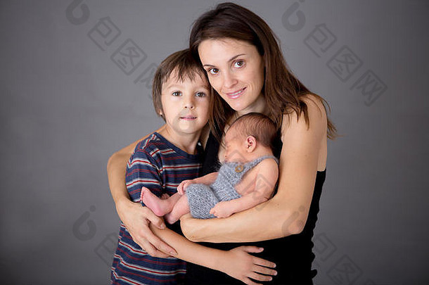 两个男孩和他们的妈妈在灰色背景下亲吻和拥抱新生婴儿的全家福，温柔，关爱。妇女和儿童肖像