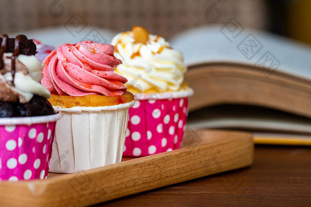 五颜六色的纸杯蛋糕放在木托盘上，打开木桌上的书。生活方式和放松的概念。