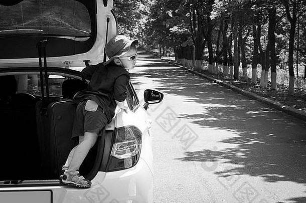 黑白照片中，一个顽皮的男孩站在一辆白色掀背车的敞篷卡车上，戴着现代太阳镜和帽子向外张望。