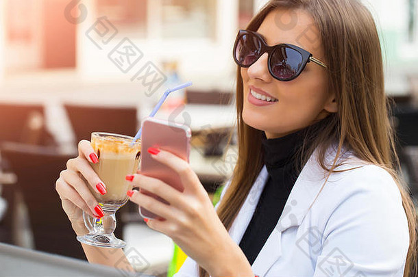 戴着太阳镜的时尚快乐女人在咖啡馆里用智能手机，手里拿着一杯现成的饮料。闭合