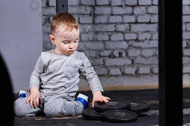 穿着运动服的可爱小男孩坐在健身房的地板上，看着砖墙上的杠铃。