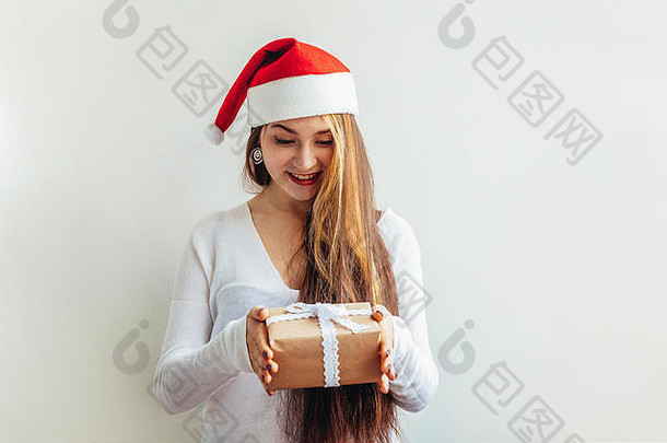 戴着红色圣诞老人帽、留着长发的漂亮女孩，手拿着礼品盒，孤立地放在白色背景上，看上去既高兴又兴奋。年轻女子肖像，真实情感。圣诞快乐，新年快乐