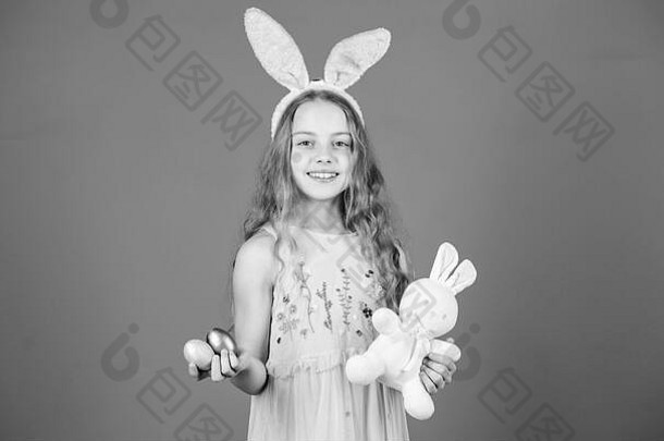 复活节快乐。戴着复活节兔子耳朵的快乐小女孩。戴着兔子头带的小女孩，手里拿着彩蛋和玩具。可爱的小女孩和可爱的填充兔子玩具。复活节兔子是复活节的象征。