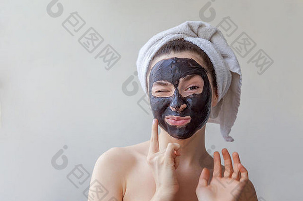 一位面带微笑的黑发女子的美丽肖像，头戴毛巾，在白色背景上脸上涂上黑色滋养面膜。护肤洁面spa放松化妆品概念