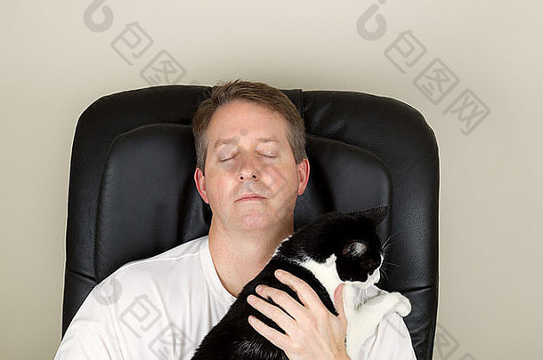 照片中，一名成年男子穿着白色衬衫，在<strong>按摩椅</strong>上放松，眼睛闭着，双手捧着黑白相间的猫