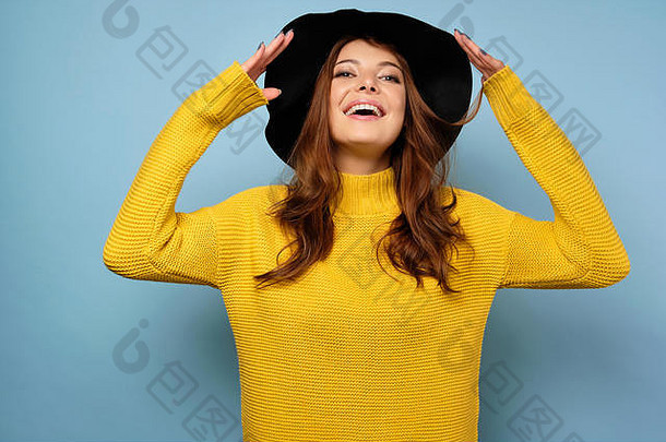 一个黑发女人穿着黄色毛衣站在蓝色的背景上，微笑着把头往后仰，黑色帽子的边缘抬起