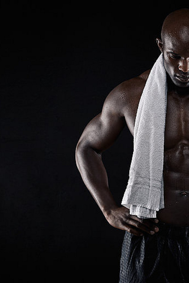 工作室拍摄了一个赤膊肌肉发达的男人，手放在臀部，手拿毛巾向下看，背景为黑色。健康的非洲男人。