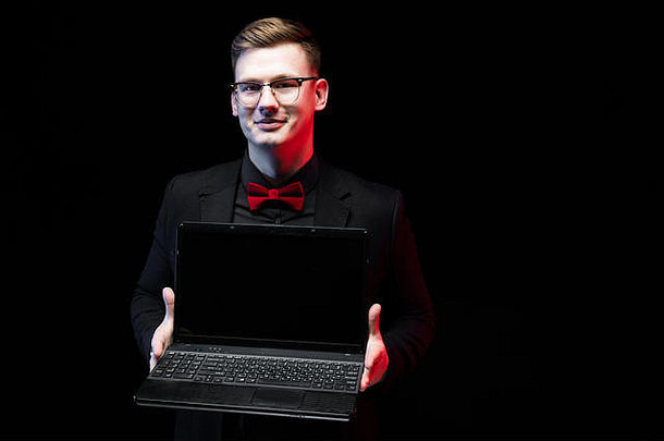自信、英俊、雄心勃勃、快乐、优雅、负责任的商人的肖像，在黑色背景下向客户提供笔记本电脑