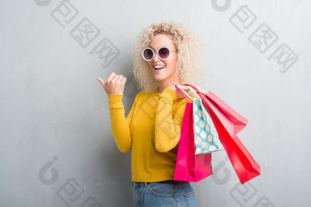 一位年轻的金发女郎站在灰蒙蒙的背景上，手里拿着购物袋，指着卖场，拇指朝上，脸上带着幸福的笑容