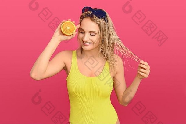 一个快乐健康的年轻女子，拿着一个切成两半的新鲜多汁的橙子放在眼睛上，在粉色的画室背景下，脸上带着友好的微笑