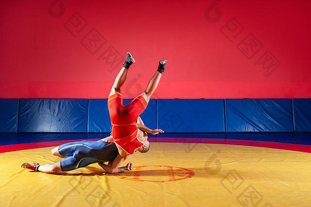 两名身穿蓝色和红色摔跤紧身衣的强壮摔跤手正在体育馆的黄色摔跤地毯上进行摔跤和超级摔跤。年轻人