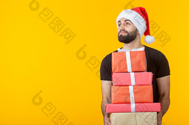 戴着圣诞老人帽子、留着胡子、心怀不满的年轻人拿着五个礼盒，在黄色背景上摆着姿势，并留有文字空间。礼物和礼物的概念