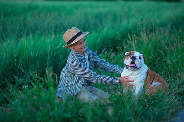 小男孩和他的英国斗牛犬站在绿色的黑麦草地上。Handsom kid穿着时髦的衣服和生锈的衣服与好友合影