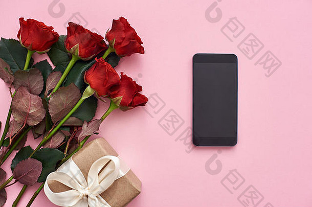 庆祝他们爱情的日子。粉红色背景下的新鲜红玫瑰、礼盒和黑色手机俯视图。