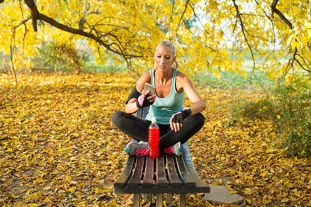 漂亮的金发女运动员用智能手机喝冷饮提神。坐在公园的长椅上。健康生活方式理念。