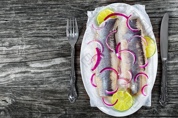 在一张古老的乡村餐桌上，用刀叉在椭圆形盘子上的羊皮纸上放着洋葱和酸橙片的鲱鱼鱼片，俯视图