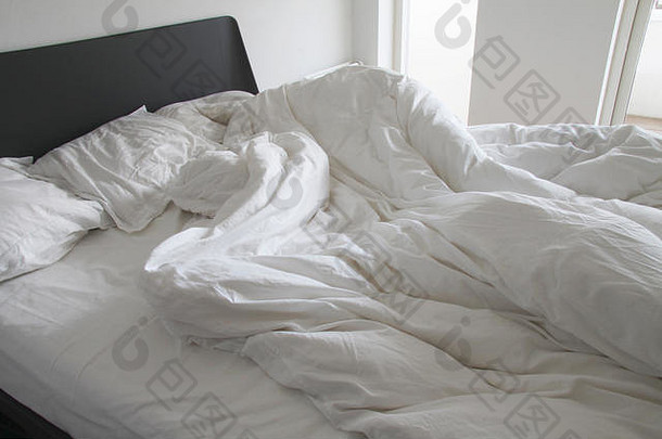 在一间白色卧室里，床上凌乱的白色床单和枕头上有皱纹