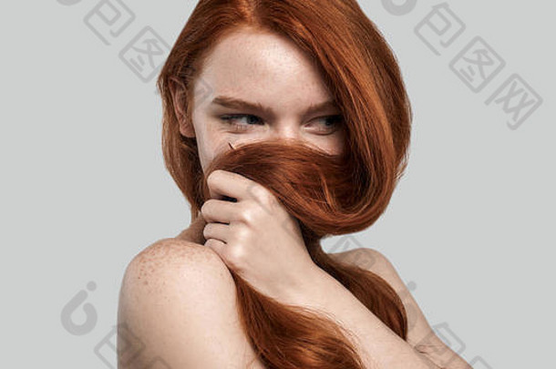 摄影棚拍摄的年轻漂亮的红发女子站在灰色背景下玩弄她的长发。美女肖像